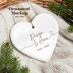 May include: A white heart-shaped ornament with the text "Design Is Here" and "PSD + PNG" in brown script. The ornament hangs from a gold string and rests on a white knitted surface with pine branches.