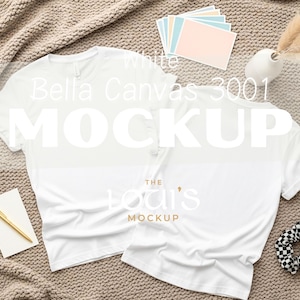 K&ouml;nnte beinhalten: Zwei wei&szlig;e Bella Canvas 3001 T-Shirts werden auf einer strukturierten beigefarbenen Oberfl&auml;che pr&auml;sentiert. Die Shirts sind so angeordnet, dass eines nach vorne und das andere gedreht ist, um die R&uuml;ckseite zu zeigen. Zus&auml;tzliche Artikel sind ein goldener Stift, Notizkarten, eine Vase mit Trockenblumen und ein schwarz-wei&szlig; kariertes Haargummi.