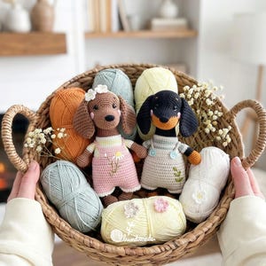 May include: A woven basket filled with crochet dachshund dogs, yarn, and flowers. One dog is brown with a pink dress, the other is black and tan with a cream dress. The yarn is in various colors, including orange, blue, and yellow.