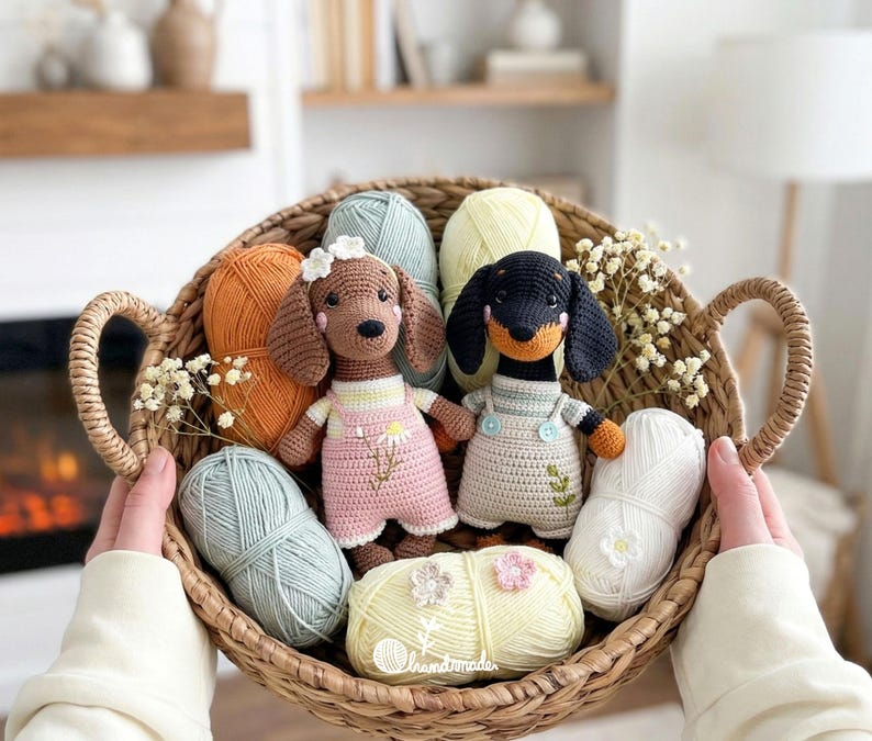 May include: A woven basket filled with crochet dachshund dogs, yarn, and flowers. One dog is brown with a pink dress, the other is black and tan with a cream dress. The yarn is in various colors, including orange, blue, and yellow.
