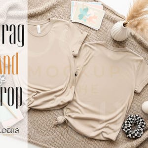 K&ouml;nnte beinhalten: Zwei beige T-Shirts werden auf einer neutralfarbenen Decke pr&auml;sentiert, die sowohl die Vorder- als auch die R&uuml;ckseite zeigen. Die Shirts sind mit hochgekrempelten &Auml;rmeln und einem geknoteten Saum versehen. Das Bild enth&auml;lt eine kleine Vase mit Trockenblumen, Schreibwaren und ein schwarz-wei&szlig;es Scrunchie.