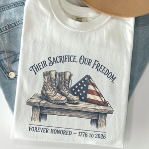 May include: White t-shirt with the text "Their Sacrifice. Our Freedom." and an illustration of combat boots and a folded American flag. The text "Forever Honored - 1776 to 2026" is printed below the illustration.