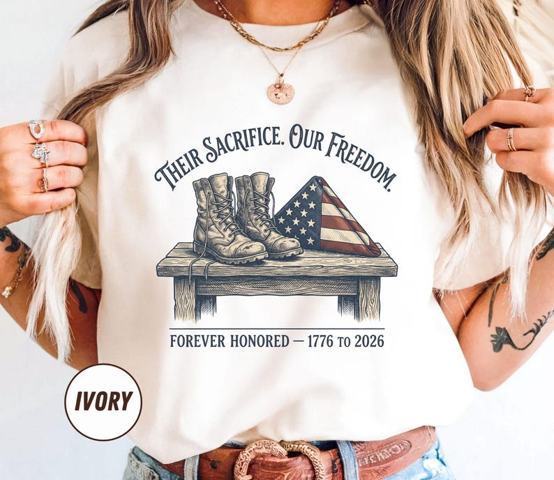 May include: Ivory-colored t-shirt featuring a graphic of combat boots, a folded American flag, and the text "Their Sacrifice. Our Freedom. Forever Honored - 1776 to 2026." The shirt also has the word "IVORY" printed on it.