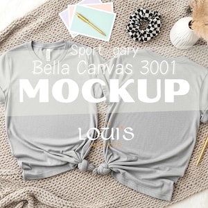 K&ouml;nnte beinhalten: Zwei hellgraue Bella Canvas 3001 T-Shirts werden auf einer strukturierten Oberfl&auml;che pr&auml;sentiert. Die Hemden sind unten geknotet. Das Wort "MOCKUP" ist prominent auf den Hemden zu sehen. Zus&auml;tzliche Artikel sind Schreibwaren, ein Scrunchie und eine Vase.