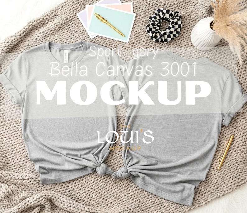 K&ouml;nnte beinhalten: Zwei hellgraue Bella Canvas 3001 T-Shirts werden auf einer strukturierten Oberfl&auml;che pr&auml;sentiert. Die Hemden sind unten geknotet. Das Wort "MOCKUP" ist prominent auf den Hemden zu sehen. Zus&auml;tzliche Artikel sind Schreibwaren, ein Scrunchie und eine Vase.