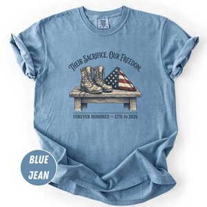 May include: Blue jean-colored t-shirt with a graphic design. The design features a pair of combat boots, a folded American flag, and the text "Their Sacrifice. Our Freedom. Forever Honored - 1776 to 2026."
