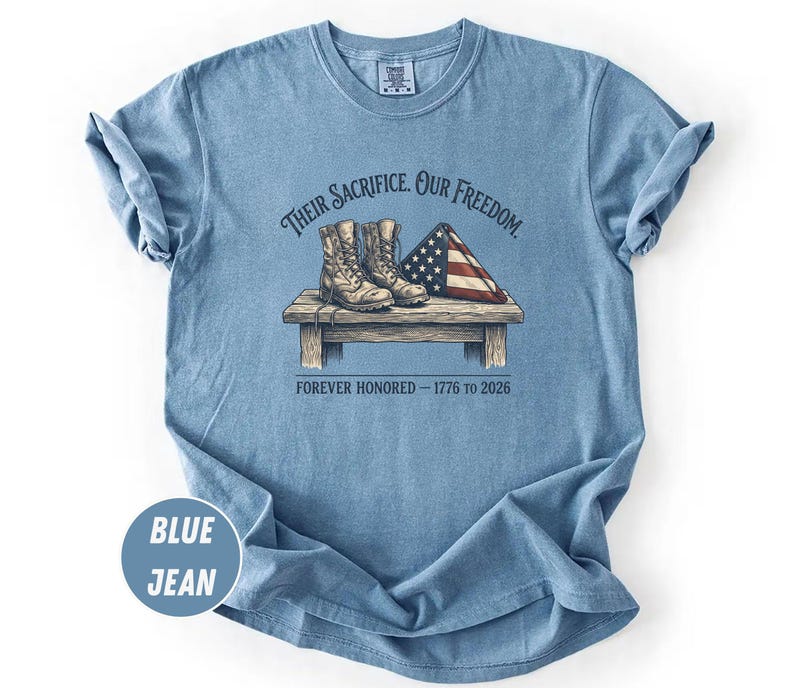 May include: Blue jean-colored t-shirt with a graphic design. The design features a pair of combat boots, a folded American flag, and the text "Their Sacrifice. Our Freedom. Forever Honored - 1776 to 2026."
