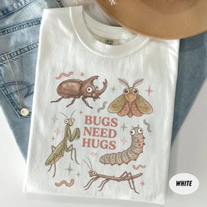May include: A white t-shirt featuring a graphic print of various insects including a beetle, moth, praying mantis, caterpillar, and stick insect. The text BUGS NEED HUGS is printed in the center in a brown font, surrounded by small star icons.