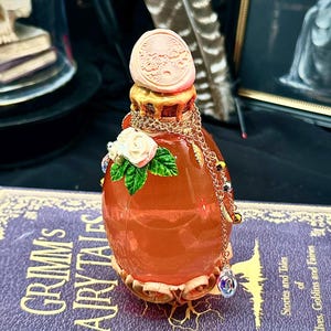 May include: An orange glass potion bottle with a decorative stopper and gold chain, resting on a purple book titled "Grimm's Fairy Tales." The bottle is adorned with a white rose and jewels. The book's cover features gold lettering.