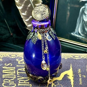 May include: A dark blue, decorative glass bottle with a detailed gold-colored stopper. The bottle is adorned with gold chains, leaf accents, and a dangling crystal. The bottle sits on a book titled "Grimm's Fairy Tales".
