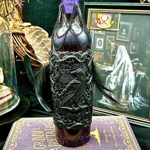 May include: A decorative bottle with a black raven design, black feathers, and a purple neck. The bottle is embellished with a chain and sits on a purple book titled "Grimm's Fairytales". The bottle's top is sealed with black wax.