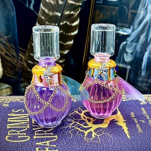 May include: Two decorative glass bottles filled with purple liquid, adorned with gold accents, chains, and fairy wings. The bottles sit on a book with the title "Grimm's Fairy Tales". The background includes a feather and framed portraits.