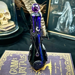 May include: A black decorative bottle with a faceted purple stopper, dripping with purple wax. A silver crescent moon and chain with a purple pendant adorn the bottle. The bottle sits on a purple book with gold lettering that reads "Grimm's Fairytales".