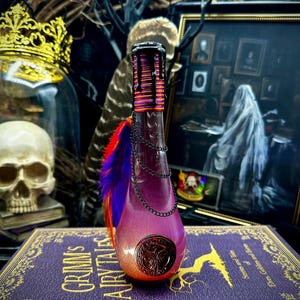 May include: A decorative, purple bottle with a chain, feathers, and a detailed stopper. The bottle is set on a purple book with gold lettering that reads "Grimm's Fairy Tales". The bottle has a metallic finish and a decorative emblem.