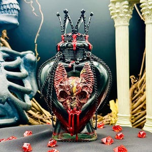 May include: A black glass bottle with a heart-shaped base and a black crown on top. The bottle is decorated with a red bat skull and dripping red paint. The bottle is surrounded by red crystals.