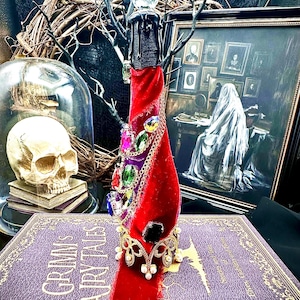 May include: A decorative bottle wrapped in red velvet, adorned with colorful jewels and a gold crown. The bottle is topped with a black candle and a crystal ball. The bottle is placed on a purple book with the title "Grimm's Fairy Tales".