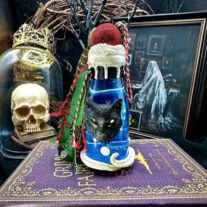 May include: A blue and white decorative bottle with a black cat face, topped with a red glitter ball and white trim. The bottle is decorated with green and red tassels and sits on a purple book with gold lettering that reads "Grimm's Fairy Tales".