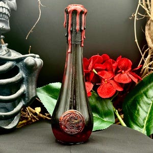 May include: A black glass bottle with a red wax seal and dripping red wax on the top. The bottle has a dark red liquid inside and a decorative design on the front.