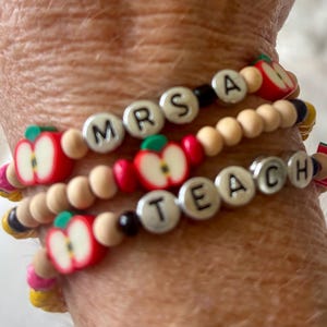 May include: A set of three beaded bracelets featuring round wooden beads, small black beads, and red apple-shaped charms. The bracelets include silver-toned letter beads spelling out the text MRS A and TEACH, arranged in a stacked design.