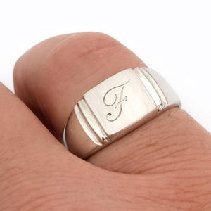 Può includere: Un anello con sigillo d'argento con una faccia rettangolare, con la lettera "F" incisa. L'anello ha una finitura lucida e viene indossato su un dito. Il design include linee verticali sui lati della faccia.