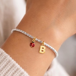 Puede incluir: Pulsera de cuentas plateadas con un dije de inicial dorada y un dije de mariquita roja y negra. La pulsera se usa en la muñeca y los dijes cuelgan libremente. El dije de inicial es la letra "B".