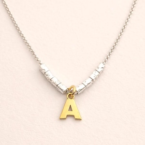 Può includere: Una collana con catena d'argento e ciondolo dorato a forma di lettera "A". La collana ha perline d'argento testurizzate e una catena delicata. Il ciondolo è di colore oro brillante.