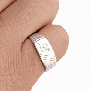 Peut inclure: Une bague en argent avec une initiale 'B' gravée sur une face rectangulaire. La bague présente un motif côtelé sur les bords supérieur et inférieur, avec des lignes diagonales sur la face. La bague est portée au doigt.