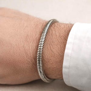 May include: A silver-colored bracelet with a ribbed design is worn on a wrist. The bracelet is made of a series of connected, curved segments. The wrist is partially visible, along with a white shirt sleeve.