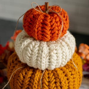 Peut inclure: Trois citrouilles tricotées empilées, aux couleurs automnales : orange, crème et orange foncé. Les citrouilles ont un motif côtelé texturé. Une cannelle et de la ficelle sont posées sur le dessus. Objet décoratif.