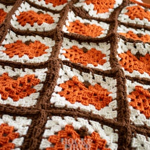 May include: A crochet blanket composed of square motifs featuring orange pumpkin designs with brown stems, surrounded by white yarn borders. The squares are joined together with thick brown yarn. The text HOOK & FIBER MADE is visible.