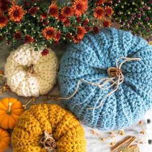 Può includere: Tre zucche all'uncinetto in bianco, blu e giallo, con bastoncini di cannella e dettagli in corda. Le zucche sono su una superficie di marmo con foglie autunnali.