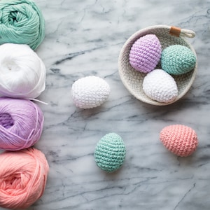 Puede incluir: Una colección de huevos de Pascua de ganchillo en una variedad de colores, incluyendo rosa, blanco, verde y morado. Los huevos están dispuestos en una cesta y sobre una superficie de mármol. También hay ovillos de lana en varios colores.