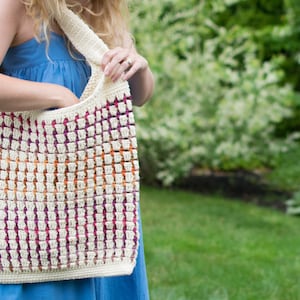 May include: A white crocheted tote bag with a colorful striped pattern in shades of purple, orange, and pink. The bag is worn over the shoulder of a person wearing a blue dress.