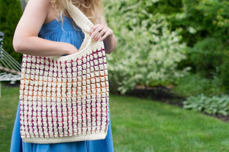 May include: A white crocheted tote bag with a colorful striped pattern in shades of purple, orange, and pink. The bag is worn over the shoulder of a person wearing a blue dress.