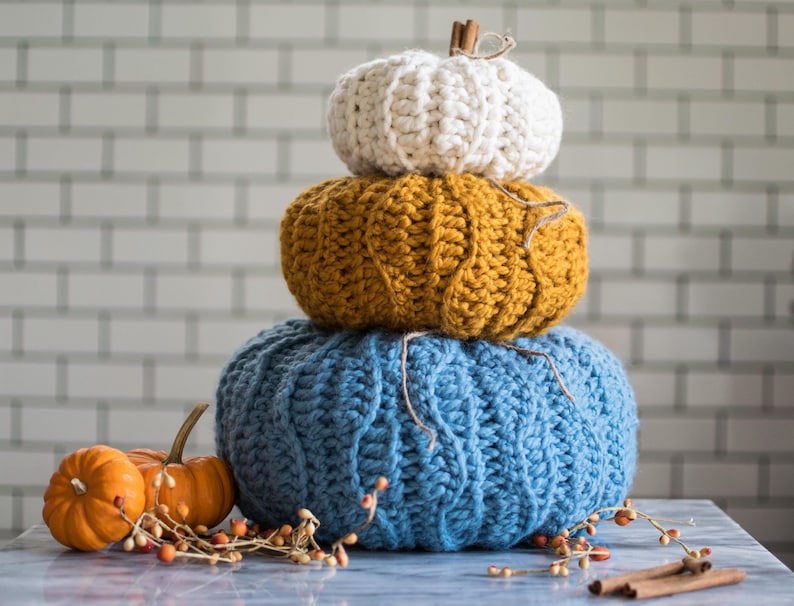 May include: A stack of three crocheted pumpkins in blue, yellow, and white. The pumpkins are arranged from largest to smallest, with the blue pumpkin on the bottom, the yellow pumpkin in the middle, and the white pumpkin on top. The pumpkins are sitting on a white marble surface.