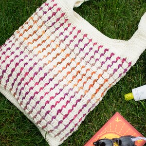 May include: A crocheted tote bag with a white base and stripes of pink, orange, and purple. The bag is lying on green grass with a book, sunglasses, a towel, and sunscreen.