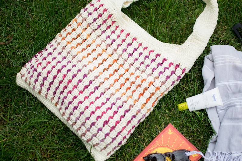 May include: A crocheted tote bag with a white base and stripes of pink, orange, and purple. The bag is lying on green grass with a book, sunglasses, a towel, and sunscreen.