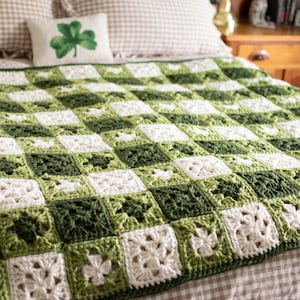 以下が含まれることがあります： 白と緑の色合いで市松模様に編まれた手作りのかぎ針編みブランケット。ベッドにはクローバーの枕が置かれています。ブランケットは居心地の良いインテリアアイテムです。