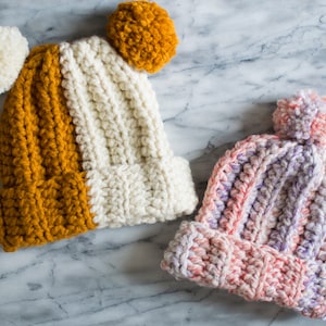 Puede incluir: Dos gorros de punto con pompones. Un gorro es blanco y amarillo mostaza, el otro es rosa y morado.