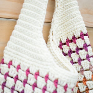 May include: A white crocheted tote bag with pink, orange, and brown accents. The bag has a wide, flat base and two long handles.