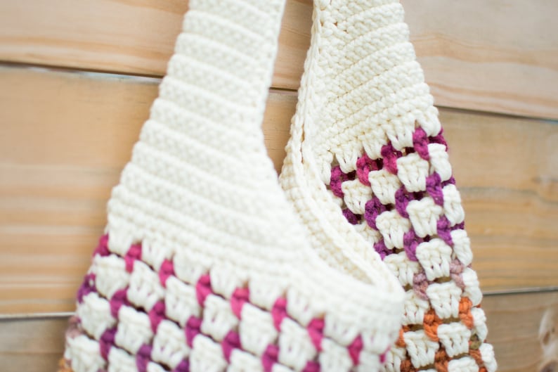 May include: A white crocheted tote bag with pink, orange, and brown accents. The bag has a wide, flat base and two long handles.