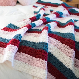 Op de afbeelding: Een gestreept gehaakt deken in tinten roze, wit, rood en blauw. Het deken is gevouwen en gedrapeerd over een wit bed.