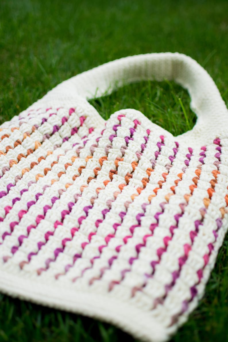 May include: A white crocheted tote bag with pink, orange, and brown stripes. The bag has a wide, flat handle.