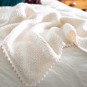 May include: A cream-coloured, hand-knitted blanket with a textured stitch pattern. The blanket is draped over a white surface, showcasing its soft, cosy appearance. The edges are finished with a decorative scalloped border, adding a touch of elegance.