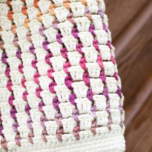 May include: A close-up of a crocheted bag with a white base and pink, orange, and purple stripes. The bag is made with a textured stitch and has a slightly uneven surface.