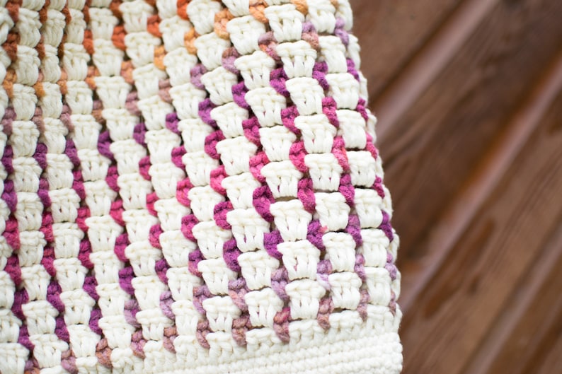 May include: A close-up of a crocheted bag with a white base and pink, orange, and purple stripes. The bag is made with a textured stitch and has a slightly uneven surface.