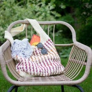 May include: A white crocheted tote bag with pink, orange, and yellow stripes. The bag is filled with a blue scarf and sunglasses. The bag is sitting on a wicker chair in a grassy area.