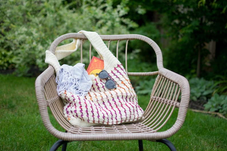 May include: A white crocheted tote bag with pink, orange, and yellow stripes. The bag is filled with a blue scarf and sunglasses. The bag is sitting on a wicker chair in a grassy area.