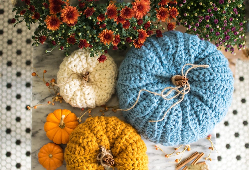 May include: Three crocheted pumpkins in white, blue, and yellow, with cinnamon sticks and twine accents. The pumpkins are sitting on a marble surface with fall foliage.