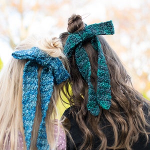 May include: Two people with long hair wearing blue crocheted hair bows. The bows are tied in a knot at the back of their heads.